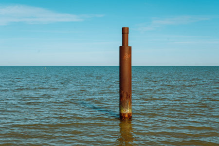 Large rusty steel construction pillar in blue sea with calm waves. Vertical abandoned building part,...