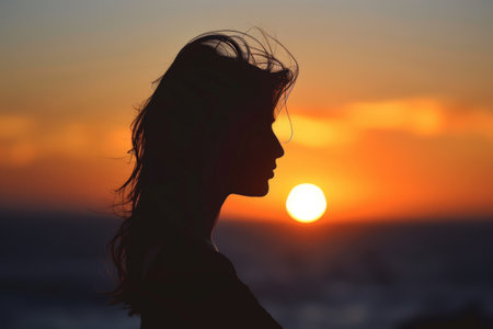 Profile silhouette of a woman against a vibrant sunset backdrop, with the glowing sun setting on the horizon.の素材