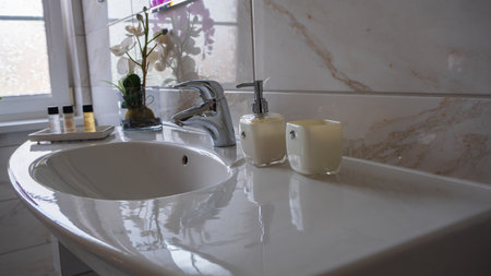 A sleek, modern bathroom sink with a faucet, soap dispenser, candle, and decorative flowers on a tiled counter.の写真素材