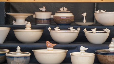 A collection of handcrafted ceramic bowls and pots adorned with bird figurines, displayed on shelves at a craft fair.の写真素材