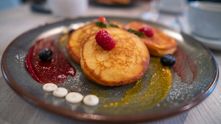 A close-up of fluffy pancakes garnished with fresh raspberries and blueberries, served with vibrant sauces on a stylish plate.の写真素材