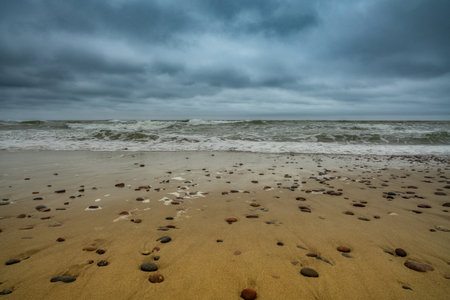 A rocky sandy beach with scattered stones and waves crashing under a moody, cloudy sky, creating a dramatic coastal scene.の写真素材