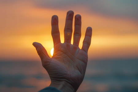 Silhouette of an open hand with fingers spread wide against a vibrant sunset sky, symbolizing hope and aspiration.の素材