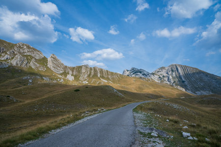 A winding road leads through grassy hills and jagged rocky peaks, set under a bright blue sky with scattered clouds.の写真素材