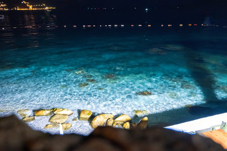 A night shot of clear turquoise waters gently hitting rocks by the shore, illuminated by nearby lights and distant reflections.の写真素材