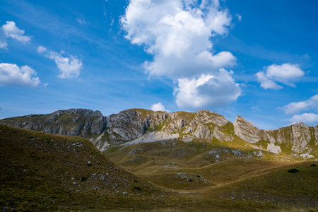 A scenic view of rugged rocky mountain peaks with grassy hills, set against a bright blue sky and scattered clouds.の写真素材