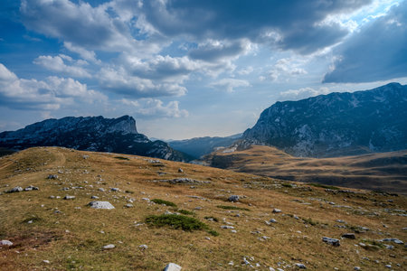 A breathtaking mountainous landscape with rocky terrain, grassy slopes, and dramatic clouds casting shadows over the distant peaks.の写真素材