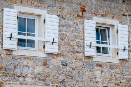 Two white shuttered windows on a rustic stone wall, blending traditional architectural elements with a charming and timeless design.の写真素材