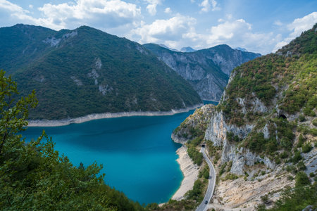 A scenic road winds through a mountain canyon alongside a vibrant blue lake, surrounded by forested cliffs and rocky terrain.の写真素材