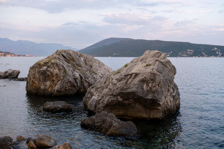 Two large rocks rest near the shore in calm water at dusk, with distant hills and a serene coastal backdrop.の写真素材
