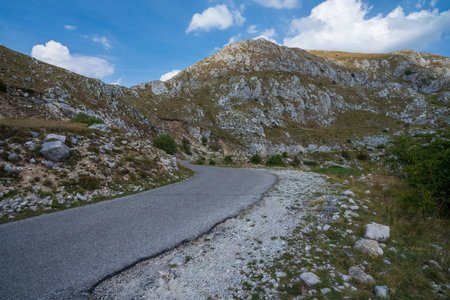 A winding road cuts through rugged, rocky mountains, surrounded by uneven terrain and sparse vegetation under a bright blue sky with scattered clouds.の写真素材