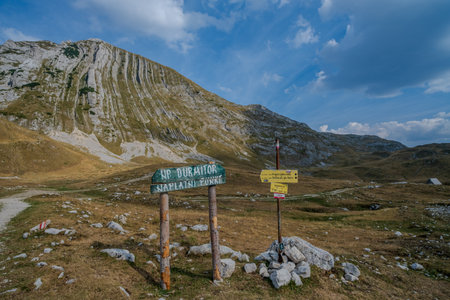 Trail signs in Durmitor National Park stand against a backdrop of rugged rocky mountains and grassy terrain under a blue sky.の写真素材