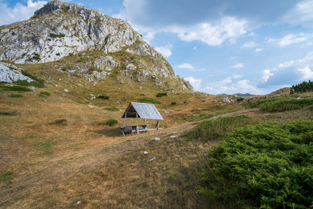 A wooden picnic shelter stands in a grassy meadow below a rocky mountain, offering a peaceful spot for outdoor relaxation.の写真素材
