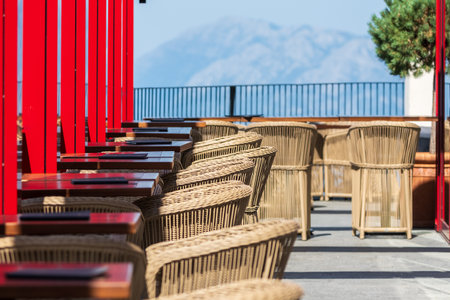 Empty outdoor cafe with wicker chairs and red tables, offering a beautiful view of distant mountains on a clear day.の写真素材