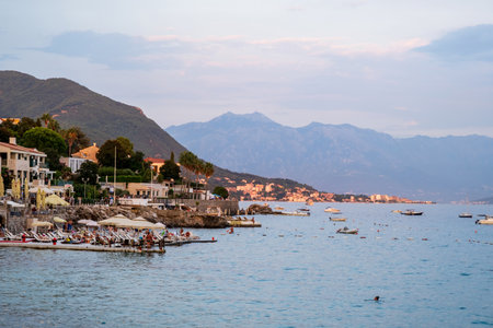 A beautiful seaside resort with sunbathers and umbrellas, overlooking calm waters and distant mountains at sunset.の写真素材