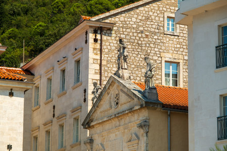 Stone statues adorn the rooftop of a historic stone building with red-tiled roofs and classical architecture, set against modern and traditional structures.の写真素材