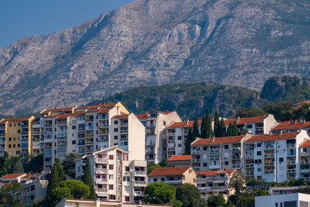 A cluster of apartment buildings with red-tiled roofs nestled at the base of a rugged mountain, surrounded by greenery in a Mediterranean town.の写真素材