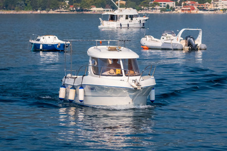 A small motorboat cruising through calm coastal waters, surrounded by other anchored boats on a sunny day near the shoreline.の写真素材