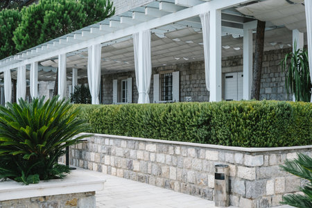 A stone building with a shaded pergola and white curtains, surrounded by lush green plants and a neatly trimmed hedge.の写真素材