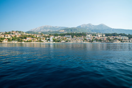 A picturesque coastal town with a backdrop of mountains and calm blue waters, featuring a blend of residential and urban buildings under a clear sky.の写真素材