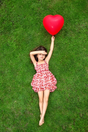 beautiful girl in color dress lying on the grass and holding a red ball in the form of heartの写真素材