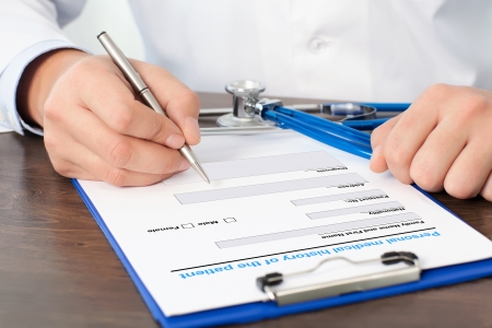 Doctor sitting at his desk with a stethoscope and writing something on a white sheetの写真素材