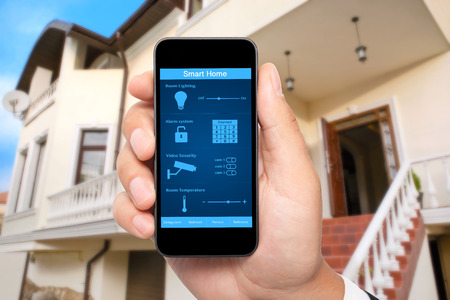 male hand hold a phone with system smart house on a screen on the background of the house male hand hold a phone with system smart house on a screen on the background of the house