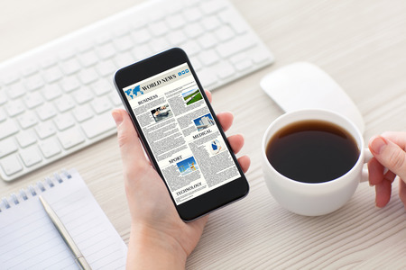 Women hands holding phone with world news site on the screen and cup of coffee in the officeの写真素材