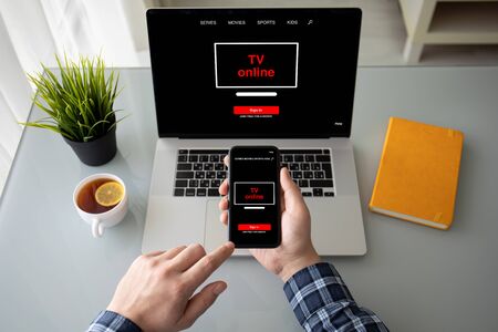 male hands holding phone over laptop with online tv application on screen at table in roomの写真素材