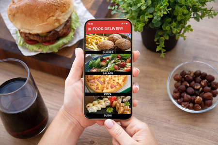 female hands holding phone with food delivery app on screen over wooden tableの写真素材