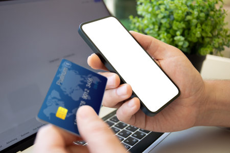 male hand holding plastic credit card and phone with isolated screen over laptop in office