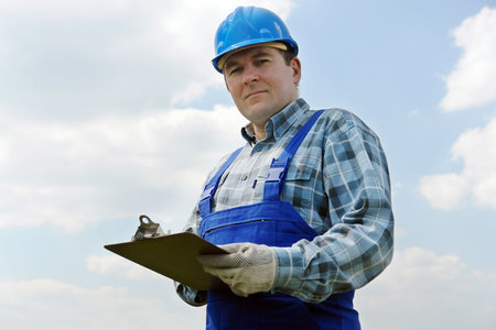 Construction site manager wearing blue helmet and overall with notepad over sky backgroundの写真素材