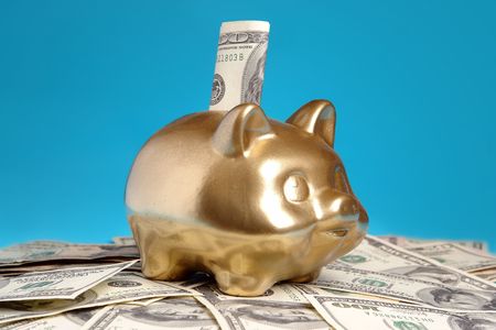 Golden piggybank with 100 US dollar bill inserted into slot placed on pile of US dollar banknotes over blue backgroundの写真素材