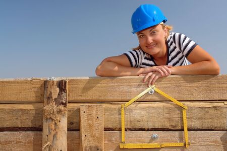 Young female wearing blue helmet posing over yellow house shaped wooden folding ruler affixed to wooden shuttering wall over blue skyの写真素材