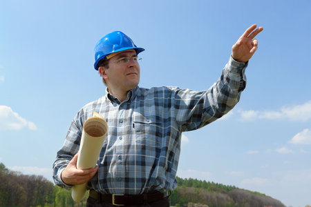 Construction site manager wearing blue helmet holding roll of building plans pointing ahead with his...