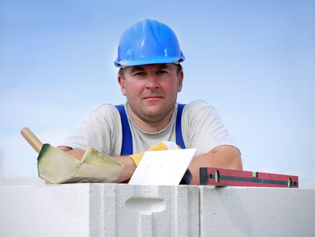 Mason wearing blue helmet posing resting on white block wall over blue sky