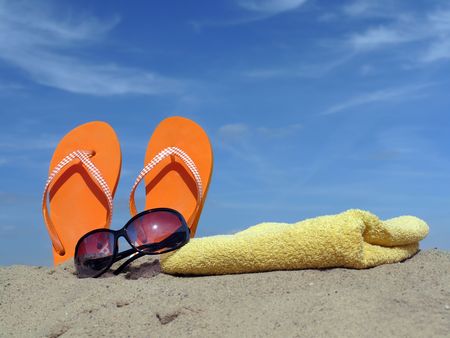 Orange flip-flops driven vertically into beach sand with sunglasses and yellow bath towel over blue skyの写真素材