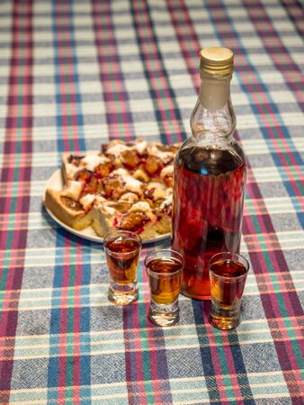 Bottle of home-made doogwood liqueur with three vodka glasses and plate of home-baked cake served on tableの写真素材