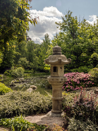 A beautifully landscaped Japanese garden featuring a traditional stone lantern, a calm pond surrounded by blooming flowers, and lush greenery under a bright skの写真素材