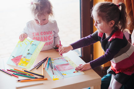 Two little girls drawing a colorful pictures of house and playing children using pencil crayons standing at table indoorsの写真素材