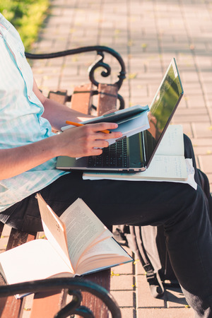 Student working on a laptop using books and notes sitting on a bench in a park. Young boy wearing a blue shirt and dark jeansの写真素材
