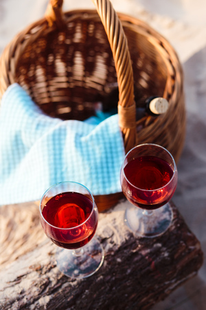 Two wine glasses with red wine standing on tree trunk, on beach, beside wicker basket with bottle of wine. View from aboveの写真素材