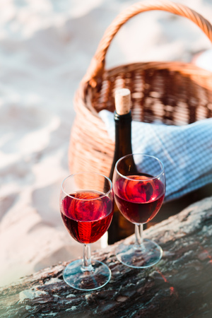 Two wine glasses with red wine standing on tree trunk, on beach, beside wicker basket with bottle of wineの写真素材