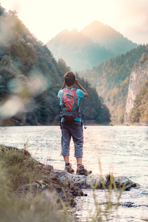 Young tourist with backpack looks through a binoculars on mountains peaks, stands on a rock over a river. Boy spends a vacation in mountains, wandering with backpack, he is wearing sports summer clothesの写真素材