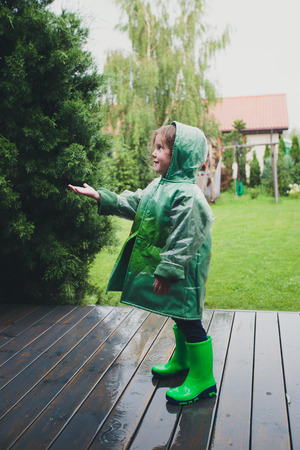 Happy smiling little girl standing on a porch catching raindrops wearing green raincoatの写真素材