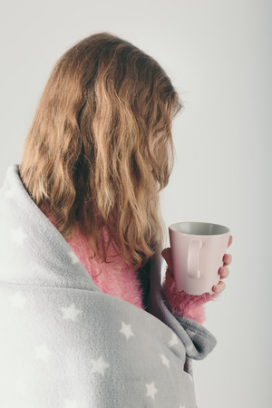 Young woman has caught cold, has day off, sick day, wrapped in blanket, holds cup of herbal tea. Profile portrait viewの写真素材