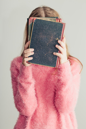 Young girl holding books in front of her face. Teenager girl wearing pink sweater. Vertical photoの写真素材
