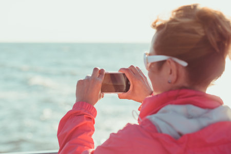 Young woman taking a photos using phone, looking at screen, standing outdoors, she is backlighted by...
