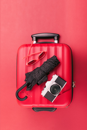 Red travel suitcase with red sunglasses, old camera, black umbrella on red background. Minimal styleの写真素材