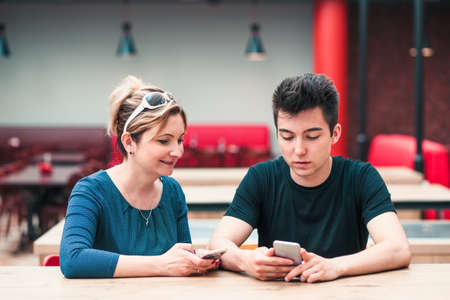 Woman and young man talking together and using mobile phones sitting by a table in cafeの写真素材